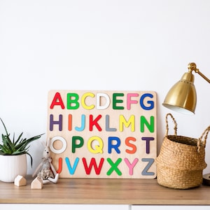May include: A wooden alphabet puzzle with colorful letters on a white background. A small stuffed bunny and a potted plant are on a wooden shelf below the puzzle.