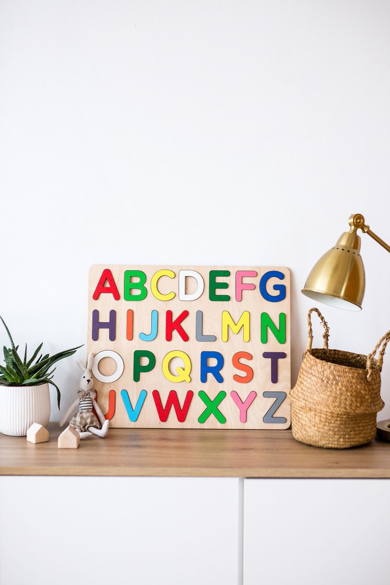 May include: A wooden alphabet puzzle with colorful letters on a white background. A small stuffed bunny and a potted plant are on a wooden shelf below the puzzle.