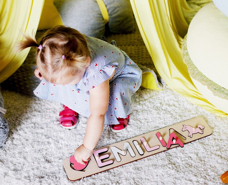 May include: A child plays with a wooden name puzzle. The puzzle is made of pink and white painted wood and spells out the name "EMILIA". The puzzle pieces are shaped like letters and a unicorn.