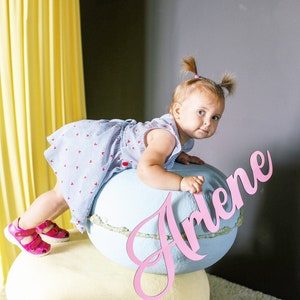 May include: A young girl in a blue and white striped dress with pink hearts sits on a large blue and white macaron with the name Arlene written in pink on the side.
