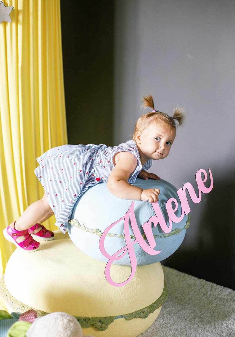 May include: A young girl in a blue and white striped dress with pink hearts sits on a large blue and white macaron with the name Arlene written in pink on the side.