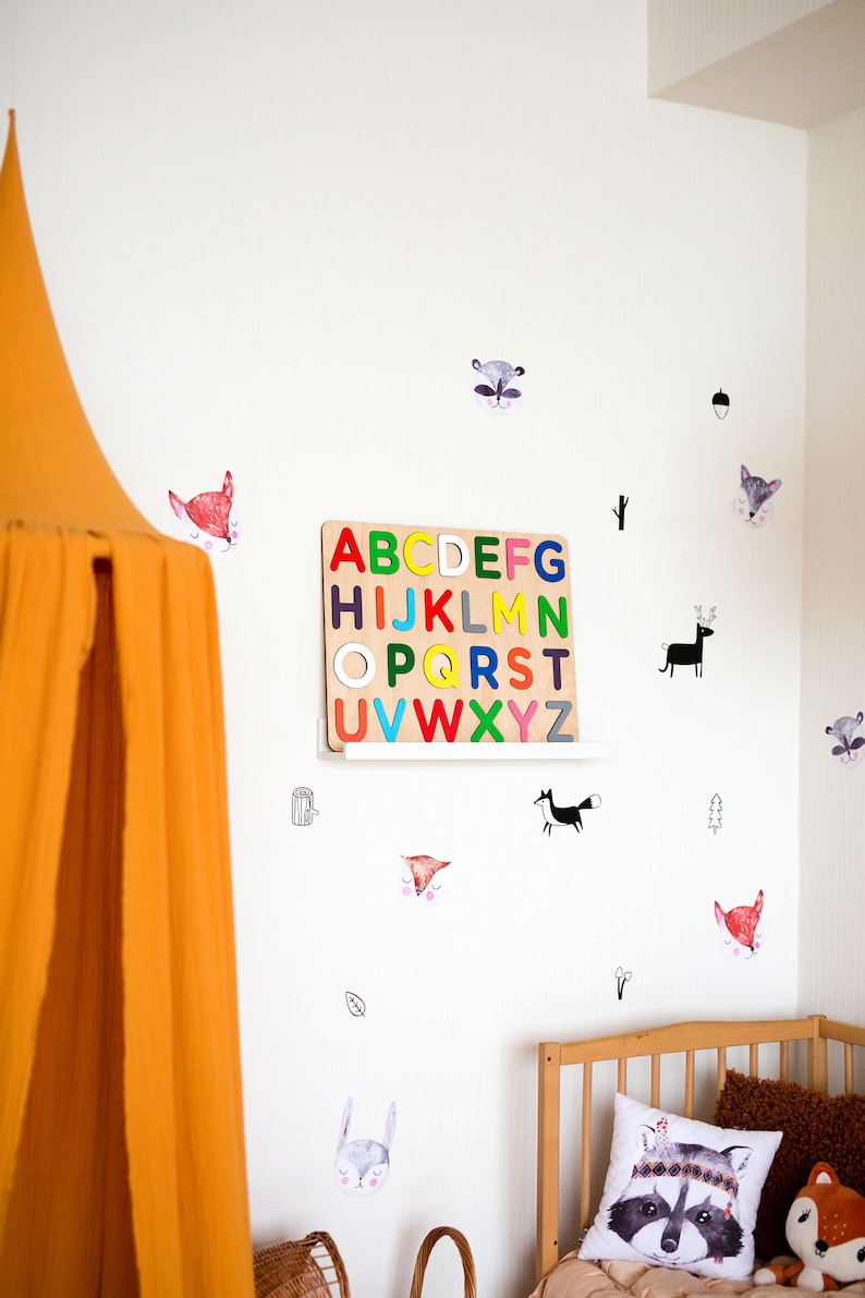 May include: A white wall with a wooden alphabet puzzle, colorful letters, and various animal stickers. The stickers include foxes, raccoons, deer, and a squirrel. There is a yellow tent canopy in the left corner of the image.