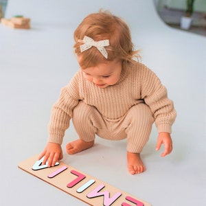 Puede incluir: Un rompecabezas de madera con el nombre "EMILIA" en letras de colores. El rompecabezas est&aacute; sobre una base de madera clara. Un ni&ntilde;o con un su&eacute;ter y pantalones beige est&aacute; jugando con el rompecabezas.