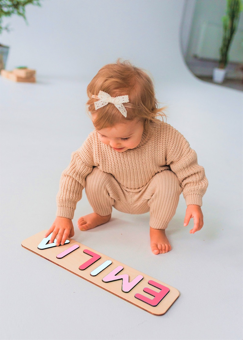 Puede incluir: Un rompecabezas de madera con el nombre "EMILIA" en letras de colores. El rompecabezas est&aacute; sobre una base de madera clara. Un ni&ntilde;o con un su&eacute;ter y pantalones beige est&aacute; jugando con el rompecabezas.
