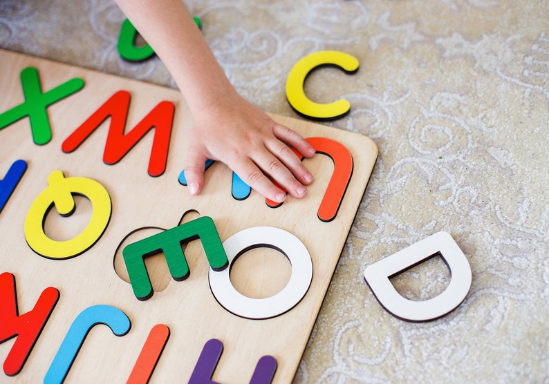May include: A child's hand reaches for a green letter 'M' from a wooden alphabet puzzle. The puzzle has colorful letters on a light wood background.