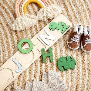 May include: Wooden puzzle with the name "JOHN" spelled out in white letters. The puzzle pieces are shaped like dinosaurs and elephants. The puzzle is on a woven rug with a rainbow and baby booties.