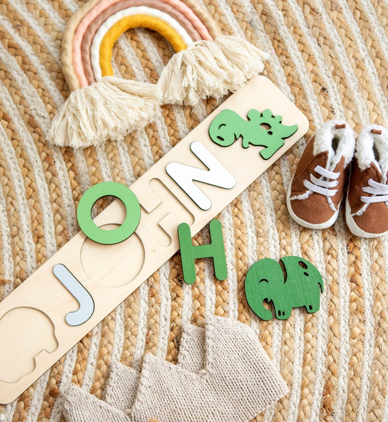 May include: Wooden puzzle with the name "JOHN" spelled out in white letters. The puzzle pieces are shaped like dinosaurs and elephants. The puzzle is on a woven rug with a rainbow and baby booties.