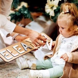 May include: A woman helps a young girl spell out the name "Eleanor" with wooden letters. The letters are painted in pastel pink, blue, and gray. The letters are on a wooden board.
