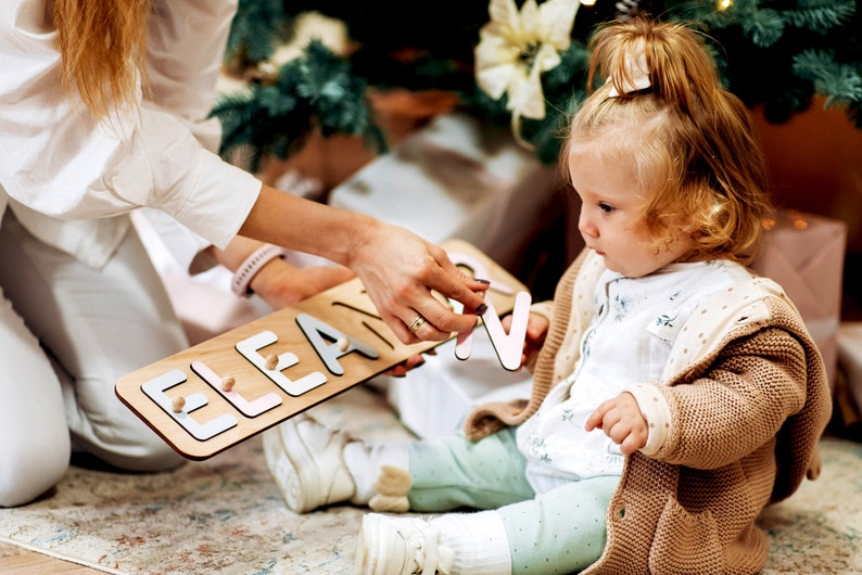 May include: A woman helps a young girl spell out the name "Eleanor" with wooden letters. The letters are painted in pastel pink, blue, and gray. The letters are on a wooden board.