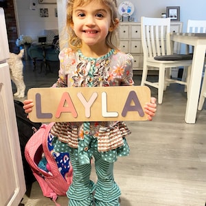 Puede incluir: Una ni&ntilde;a sostiene un rompecabezas de madera que deletrea "LAYLA" en letras coloridas. Lleva una blusa floral, pantalones a rayas turquesa y una mochila rosa es visible. El fondo incluye un interior de casa con una mesa y sillas blancas.
