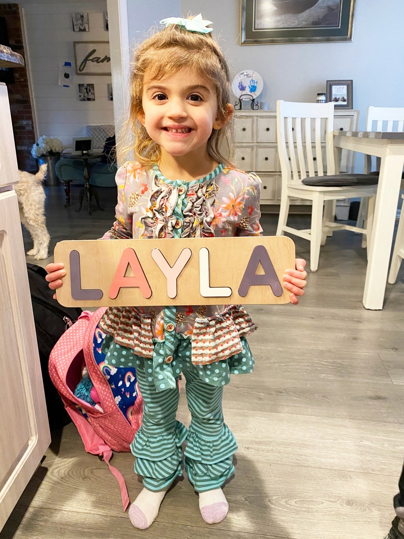 Puede incluir: Una ni&ntilde;a sostiene un rompecabezas de madera que deletrea "LAYLA" en letras coloridas. Lleva una blusa floral, pantalones a rayas turquesa y una mochila rosa es visible. El fondo incluye un interior de casa con una mesa y sillas blancas.
