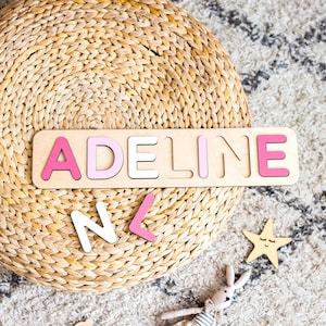 Puede incluir: Un rompecabezas de madera con el nombre "ADELINE" en letras rosas y blancas. El rompecabezas est&aacute; sobre una base de madera clara. Hay letras sueltas adicionales esparcidas cerca. Una estrella y un conejito de peluche tambi&eacute;n est&aacute;n en la imagen.