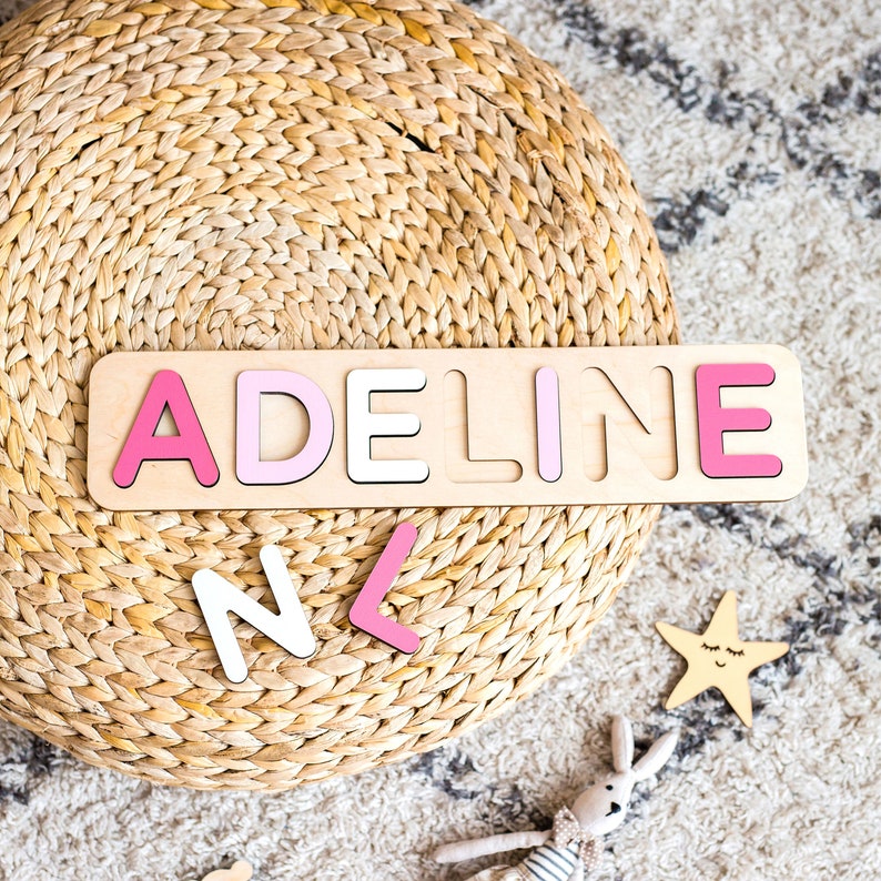 Puede incluir: Un rompecabezas de madera con el nombre "ADELINE" en letras rosas y blancas. El rompecabezas est&aacute; sobre una base de madera clara. Hay letras sueltas adicionales esparcidas cerca. Una estrella y un conejito de peluche tambi&eacute;n est&aacute;n en la imagen.