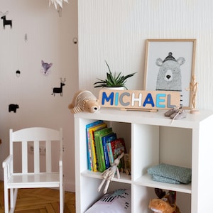 Puede incluir: Un mueble estanter&iacute;a c&uacute;bico blanco con un rompecabezas de nombre de madera que deletrea "MICHAEL" en letras azules y grises. Una ilustraci&oacute;n de oso enmarcada, un oso de peluche y una peque&ntilde;a planta se encuentran encima. Se ven libros, un conejito de peluche y un juguete de alce.