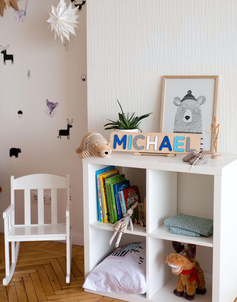 Puede incluir: Un mueble estanter&iacute;a c&uacute;bico blanco con un rompecabezas de nombre de madera que deletrea "MICHAEL" en letras azules y grises. Una ilustraci&oacute;n de oso enmarcada, un oso de peluche y una peque&ntilde;a planta se encuentran encima. Se ven libros, un conejito de peluche y un juguete de alce.