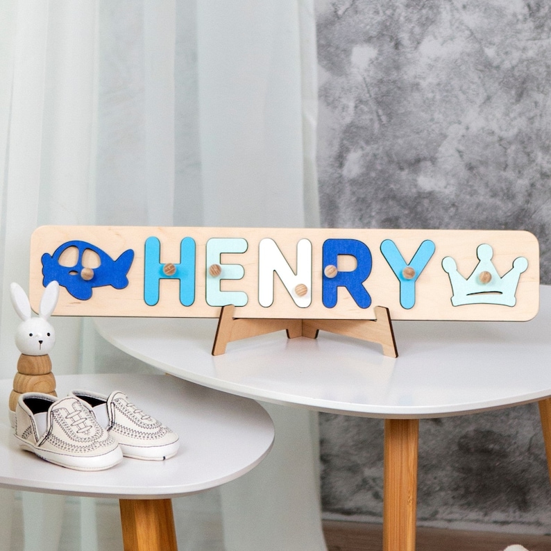 May include: A wooden name puzzle with the name "HENRY" in blue, white, and light blue letters. The puzzle has a blue airplane on the left and a blue crown on the right. The puzzle is on a wooden stand.