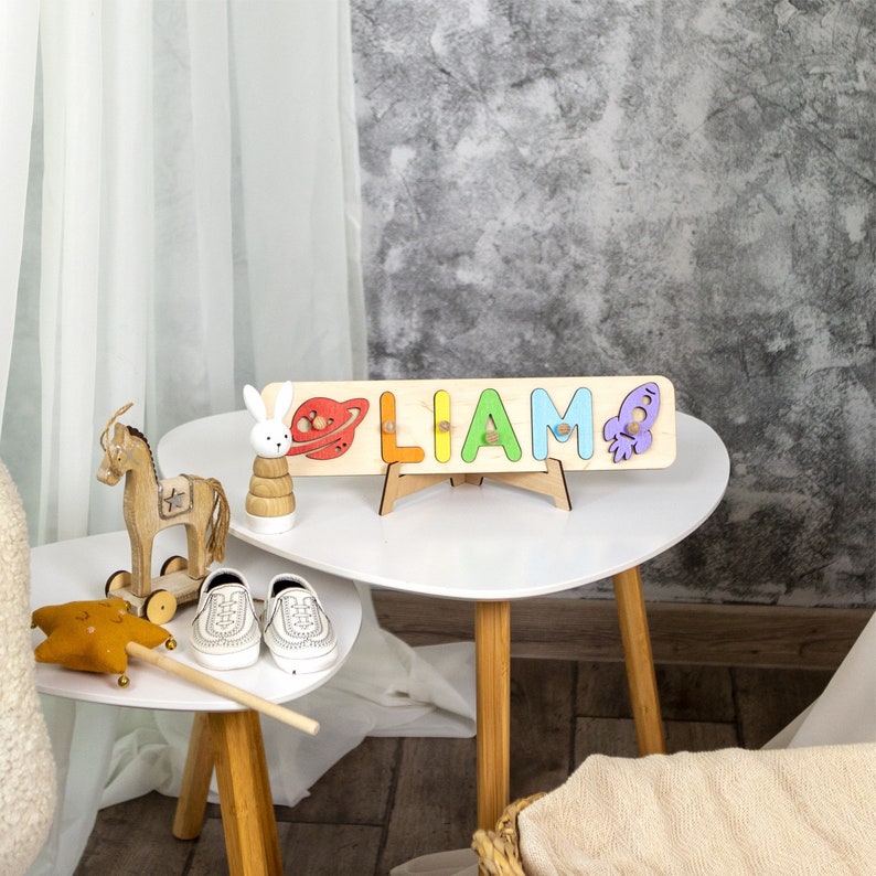 May include: A wooden name puzzle with the name "LIAM" spelled out in colorful letters. The puzzle is decorated with a rocket, a planet, and a rabbit. The puzzle is on a white stand and is sitting on a white table.