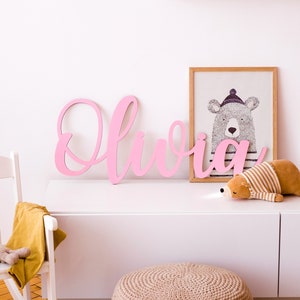 May include: Pink wooden letters spelling out the name "Olivia" on a white wall with a framed print of a bear wearing a hat above a white dresser.