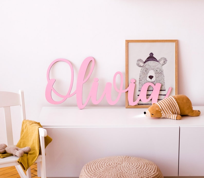 May include: Pink wooden letters spelling out the name "Olivia" on a white wall with a framed print of a bear wearing a hat above a white dresser.