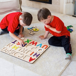 May include: Two children play with a wooden alphabet puzzle on a white rug. The puzzle has colorful letters and is laid out on the floor. The children are wearing red shirts and are focused on the puzzle.