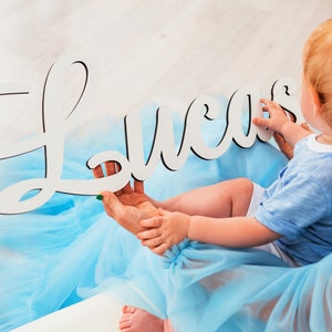 May include: A white wooden sign with the name "Lucas" spelled out in cursive. The sign is being held by a person wearing a light blue tulle dress. A baby boy is sitting on the person's lap, wearing a light blue shirt.