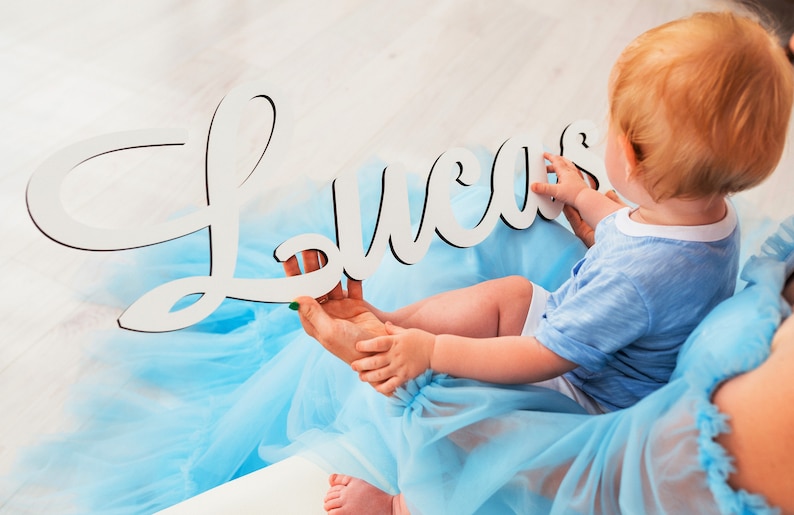 May include: A white wooden sign with the name "Lucas" spelled out in cursive. The sign is being held by a person wearing a light blue tulle dress. A baby boy is sitting on the person's lap, wearing a light blue shirt.