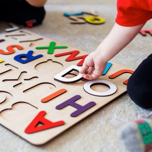 May include: A child is playing with a wooden alphabet puzzle. The puzzle has colorful letters that fit into the corresponding cutouts. The child is holding a letter D in their hand.