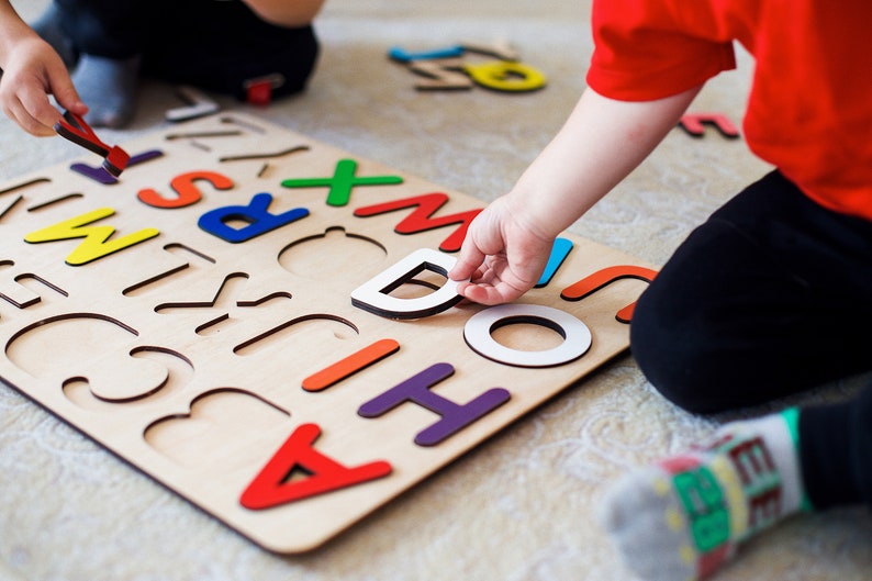 May include: A child is playing with a wooden alphabet puzzle. The puzzle has colorful letters that fit into the corresponding cutouts. The child is holding a letter D in their hand.