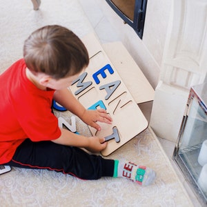 May include: A wooden puzzle board featuring cutouts for letters, with some blue and grey letter pieces inserted into the board. The board displays the letters E, A, N, M, and T in a mix of blue and grey colours, with a single T piece being held.