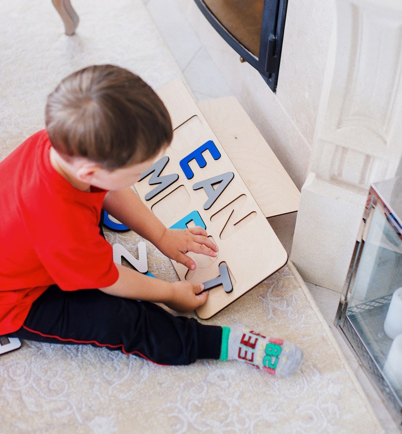 May include: A wooden puzzle board featuring cutouts for letters, with some blue and grey letter pieces inserted into the board. The board displays the letters E, A, N, M, and T in a mix of blue and grey colours, with a single T piece being held.