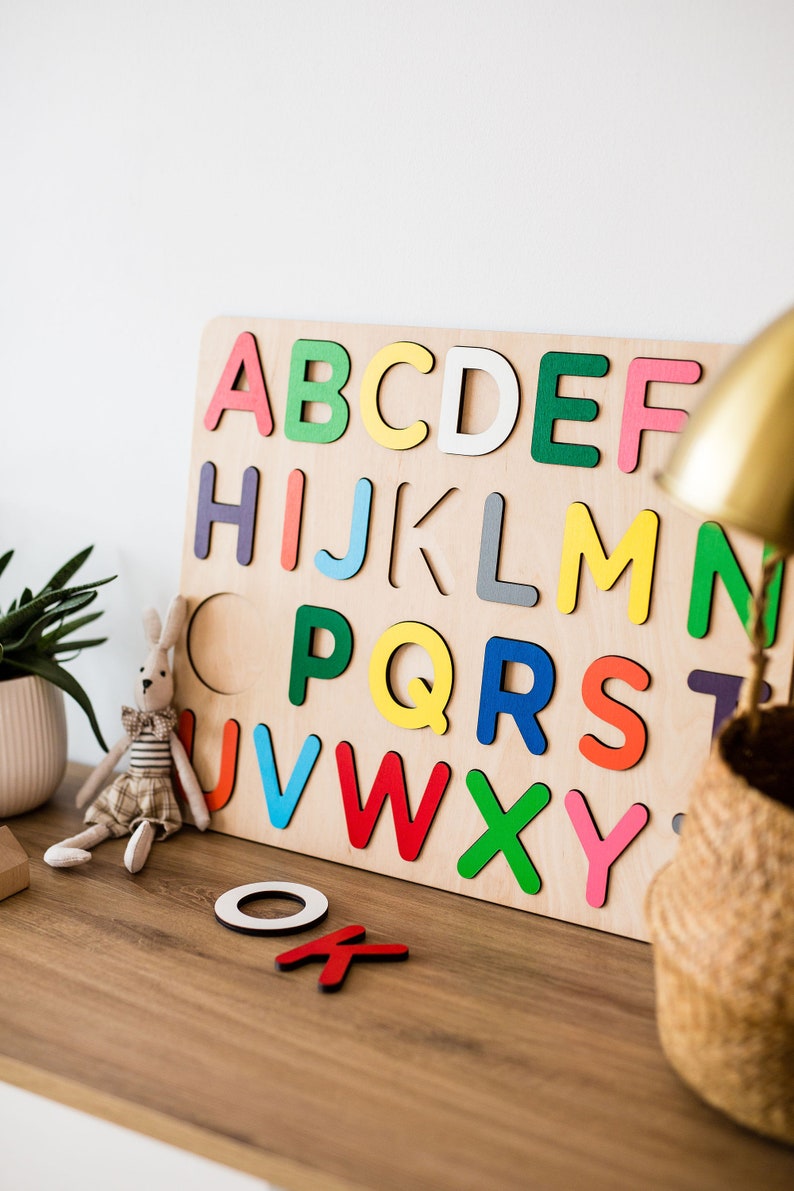 May include: A wooden alphabet puzzle with colorful letters. The letters are arranged in alphabetical order and are missing from their spaces. The puzzle is on a wooden table with a stuffed bunny and a wicker basket.
