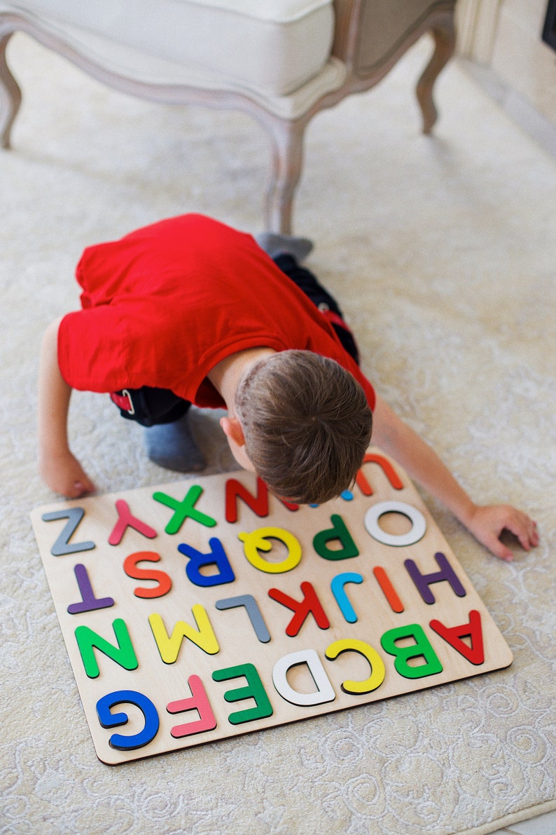 May include: A child in a red shirt is kneeling on the floor and looking at a wooden alphabet puzzle. The puzzle has colorful letters on a light brown background.