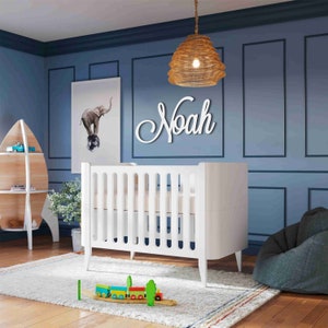 May include: White cot with a white mattress and a white wooden name sign that says "Noah" above it. A wicker pendant light hangs above the cot. There is a white bookshelf with a wooden surfboard design on the left side of the image.