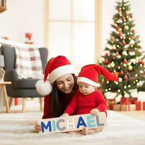 Puede incluir: Una madre y un beb&eacute; con sombreros y trajes rojos de Pap&aacute; Noel sostienen un rompecabezas de nombre de madera que deletrea "MICHAEL". El rompecabezas tiene letras azules y grises. Un &aacute;rbol de Navidad decorado est&aacute; en el fondo.