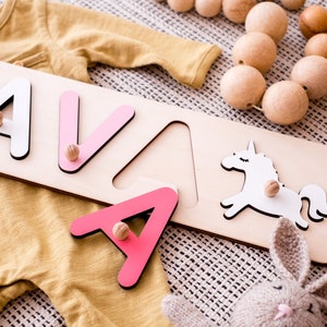 May include: A wooden name puzzle board featuring the letters A, V, and A, with a white unicorn cutout. The letters are painted in shades of white and pink, each equipped with a small wooden knob for handling. The puzzle is placed near a mustard yellow long-sleeve garment, a string of natural wooden beads, and a small knitted rabbit toy.