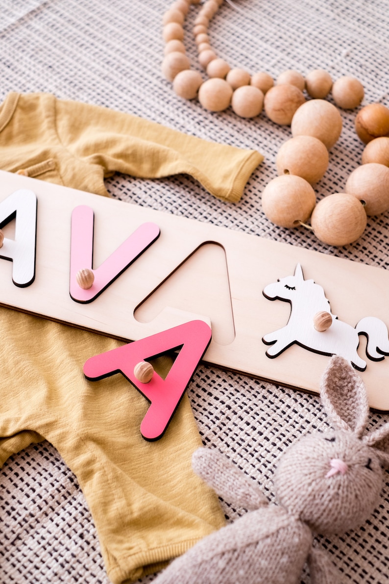 May include: A wooden name puzzle board featuring the letters A, V, and A, with a white unicorn cutout. The letters are painted in shades of white and pink, each equipped with a small wooden knob for handling. The puzzle is placed near a mustard yellow long-sleeve garment, a string of natural wooden beads, and a small knitted rabbit toy.