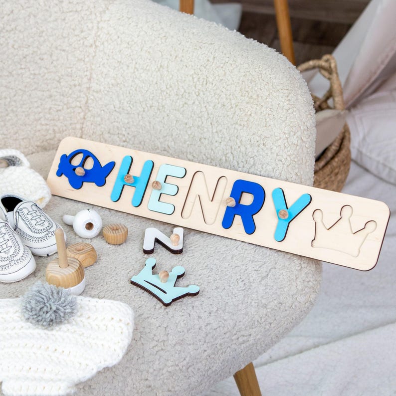 Puede incluir: Rompecabezas de madera con el nombre "HENRY" en letras azules y celeste, un avi&oacute;n azul y una corona. Hay juguetes de madera y zapatos de beb&eacute; cerca. El rompecabezas mide aproximadamente 30 cm de largo.