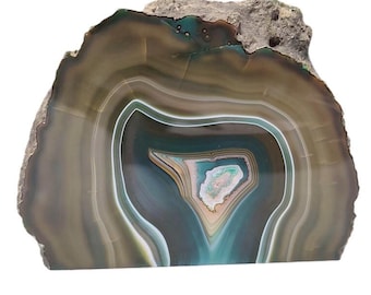 May include: A polished agate stone slice with concentric bands of brown, white, and teal. The natural stone has a rough, unpolished edge at the top and a smooth, polished face. The center features a unique, eye-like pattern.