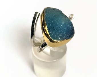 Puede incluir: Un anillo de plata y oro con una piedra de ágata azul engastada en un engaste de oro. El anillo está en un expositor blanco.