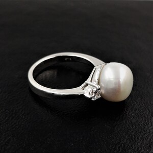 May include: A silver ring with a large white pearl and a smaller clear gemstone. The ring is set on a black background.