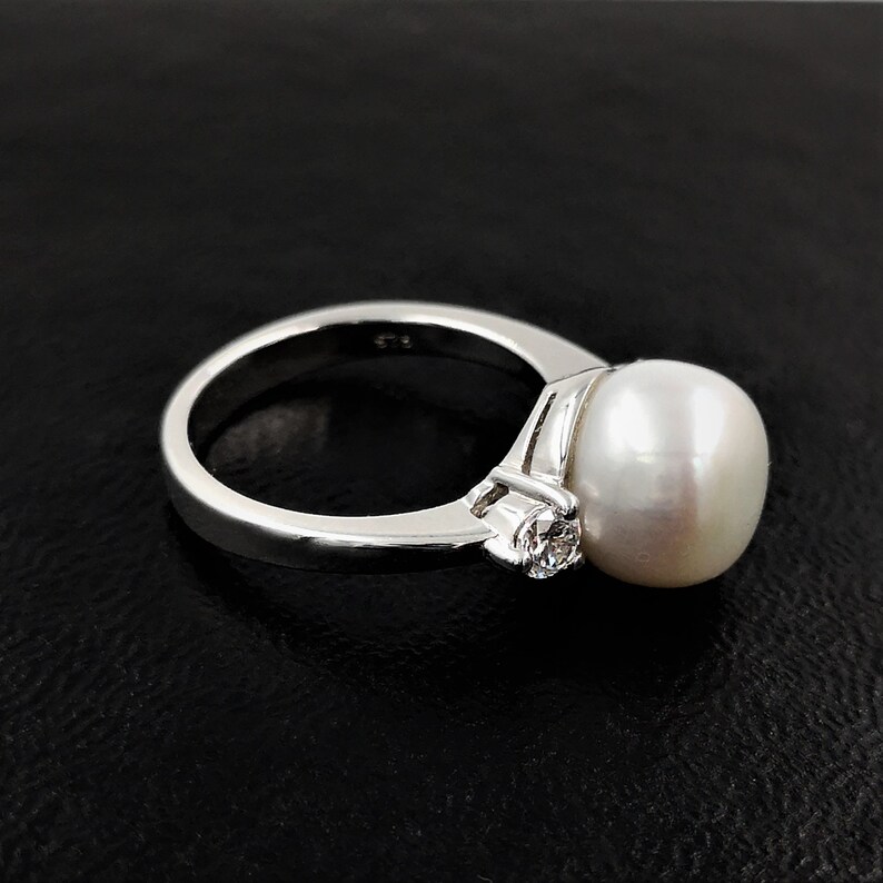 May include: A silver ring with a large white pearl and a smaller clear gemstone. The ring is set on a black background.
