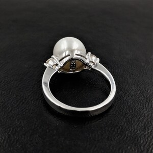May include: A silver ring with a large white pearl set in a silver and diamond setting. The ring is on a black background.