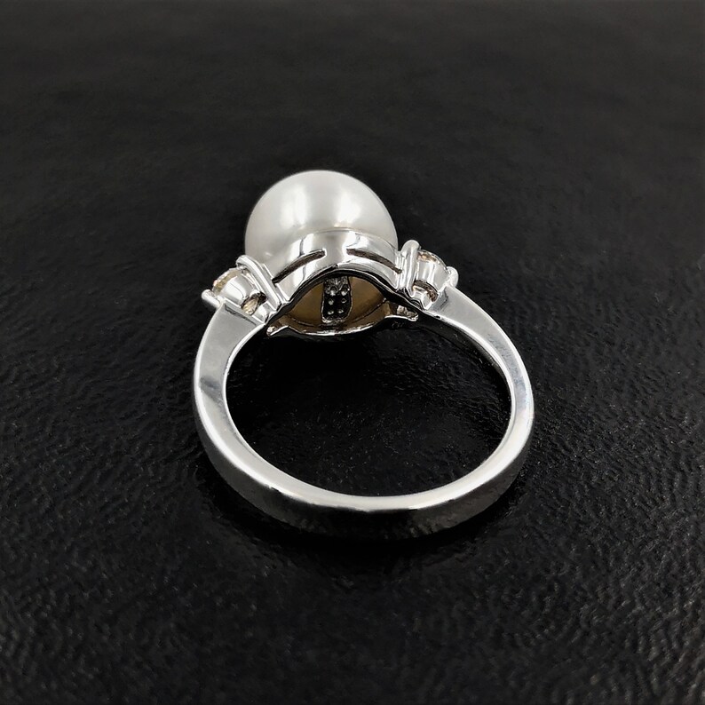 May include: A silver ring with a large white pearl set in a silver and diamond setting. The ring is on a black background.