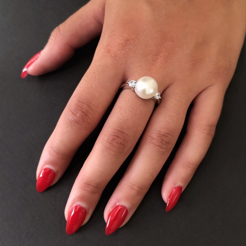 May include: A silver ring with a large white pearl and two smaller clear stones on either side of the pearl.