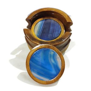Peut inclure: Ensemble de quatre sous-verres en bois avec des inserts en agate bleue. Les sous-verres sont empilés dans un support circulaire.