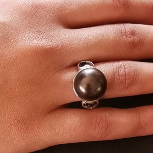 May include: A silver ring with a large, dark brown pearl set in the center. The ring has a simple design with two small loops on either side of the pearl.