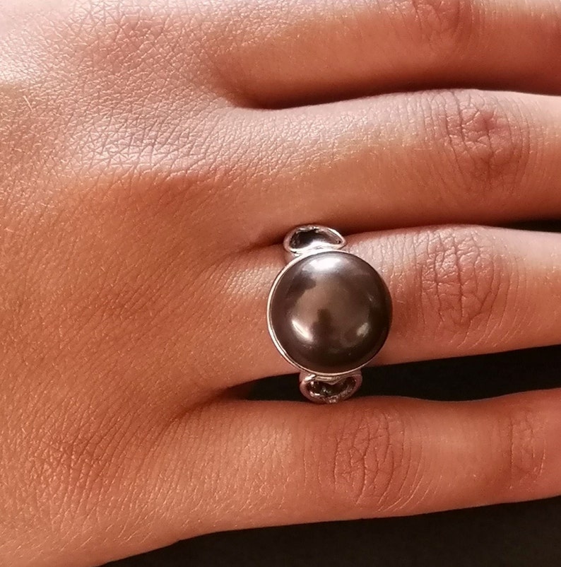 May include: A silver ring with a large, dark brown pearl set in the center. The ring has a simple design with two small loops on either side of the pearl.