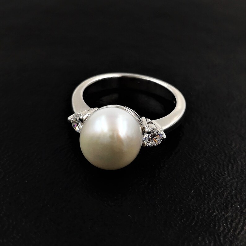 May include: A silver ring with a large white pearl and two smaller clear stones on either side.