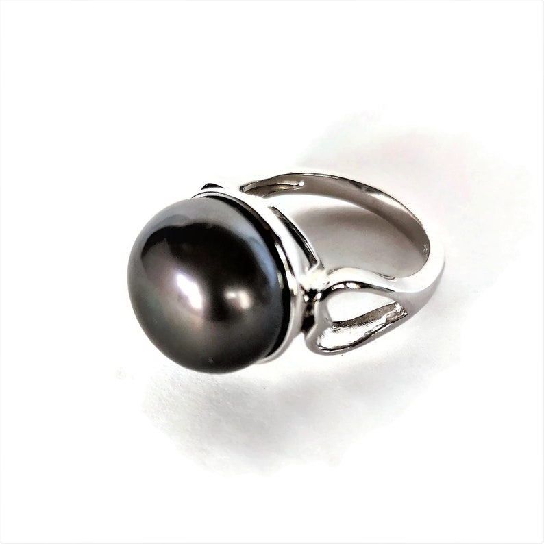 May include: A silver ring with a large, dark gray pearl set in a bezel setting. The ring has a simple, elegant design.