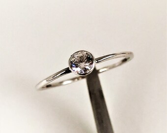 May include: A delicate silver ring featuring a single, round, clear gemstone. The gemstone is set in a bezel setting, and the band is thin and polished. The ring is displayed against a white background.